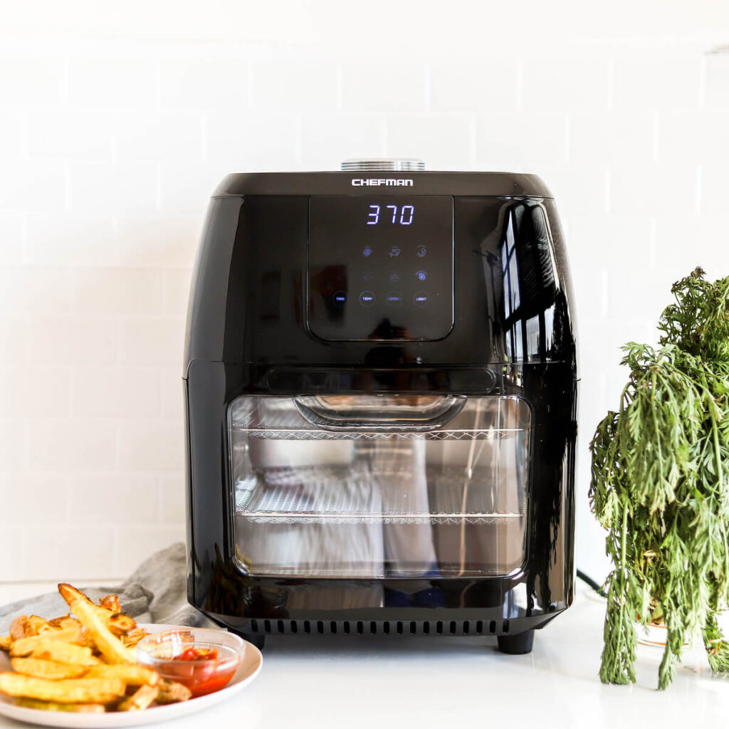 The Chefman Multi-Function Air Fryer sitting on a counter by a plate of French fries and some herbs.