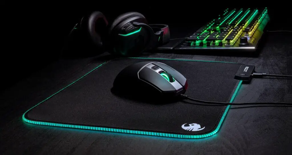 The Roccat Sense Aimo mouse pad lit up with RGB lighting in use. A mouse lays on it while it is next to a keyboard and a headset with matching RGB.