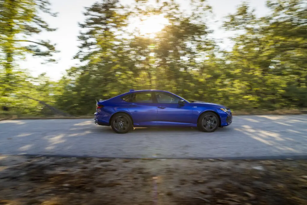 A deep blue Acura TLX driving on a road lined with trees.