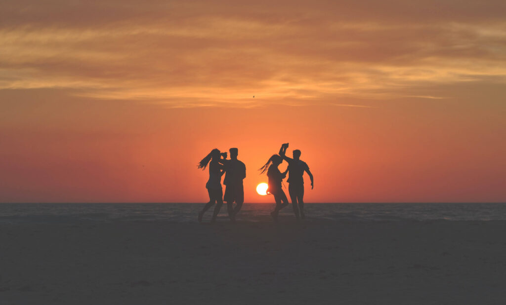Friends dancing to the music of their Fugoo 2.0 Bluetooth speakers while on the beach in a sunset.