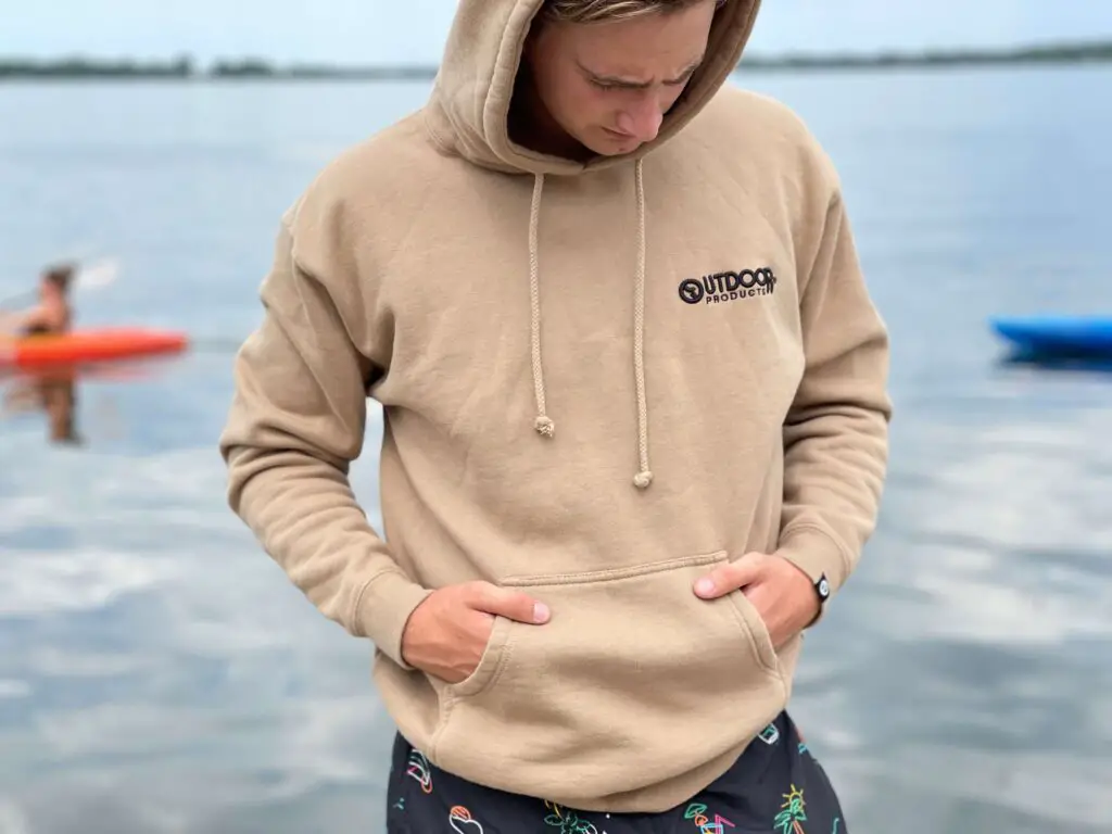 Elias wearing his '73 Originals Sandstone hoodie as he stands in by a lake.