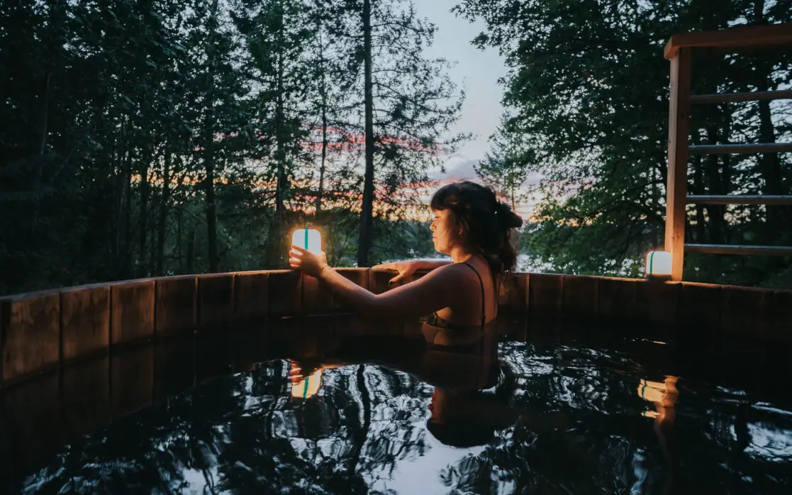 A woman in a hot tub during the sunset in a beautiful forest while she puts on her BioLite AlpenGlow 250 and 500 lumen lanterns.