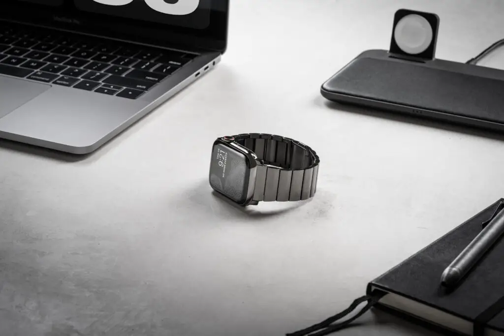 The Nomad Titanium watchband on an Apple Watch which is placed next to a MacBook and on a desk.